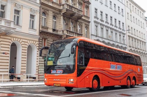 Помаранчевий автобус MAN Lion’s Coach перевізника ТрансТемпо проїжджає засніженою міською вулицею повз історичні будівлі — бронювання квитків на міжнародні маршрути через atatickets.com.
