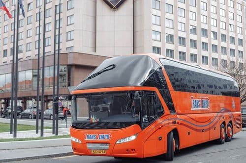 Помаранчевий лайнер Neoplan Starliner L від ТрансТемпо класу Euro-5 проїжджає повз скляний бізнес-центр — надійні пасажирські перевезення з atatickets.com.