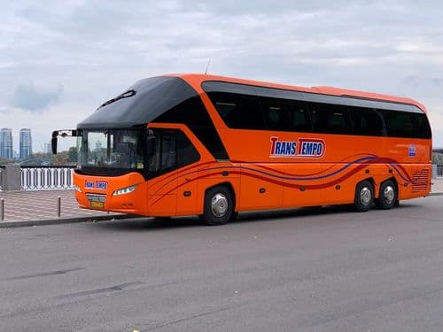 Помаранчевий автобус Neoplan Starliner перевізника TransTempo, припаркований на набережній річки з видом на міські хмарочоси вдалині — зручне бронювання квитків на міжнародні рейси через atatickets.com.